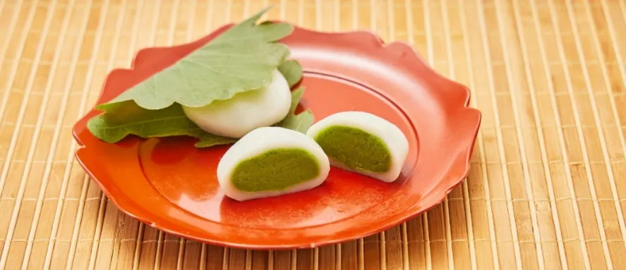 朱色の皿に盛り付けられた、鮮やかな緑の抹茶餡が覗く柏もち＜抹茶＞と、柏の葉に包まれたお餅